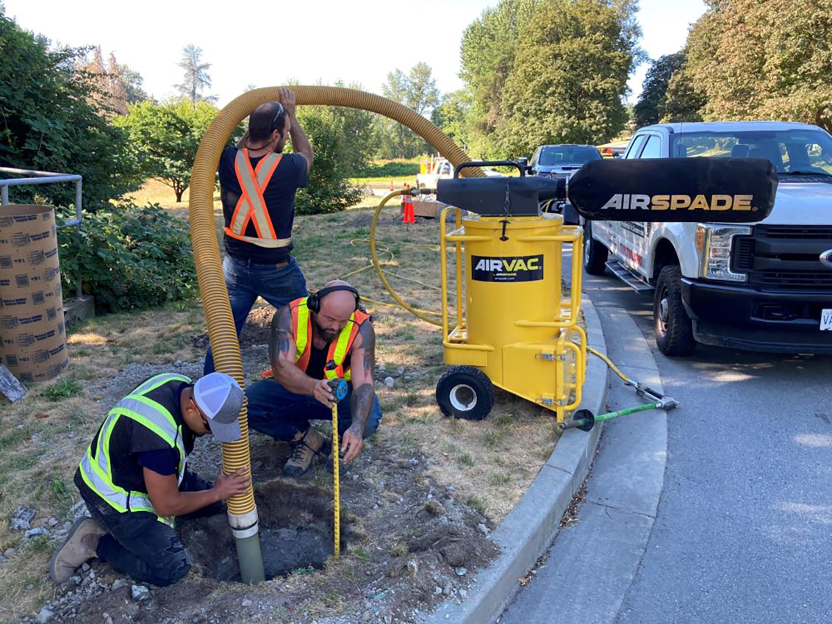3 individuals using a large vacuum type of machine in a hole dug in the ground