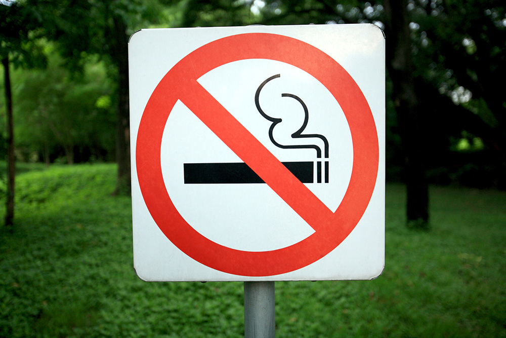 No Smoking sign with a crossed-out cigarette icon, set against a green outdoor background.