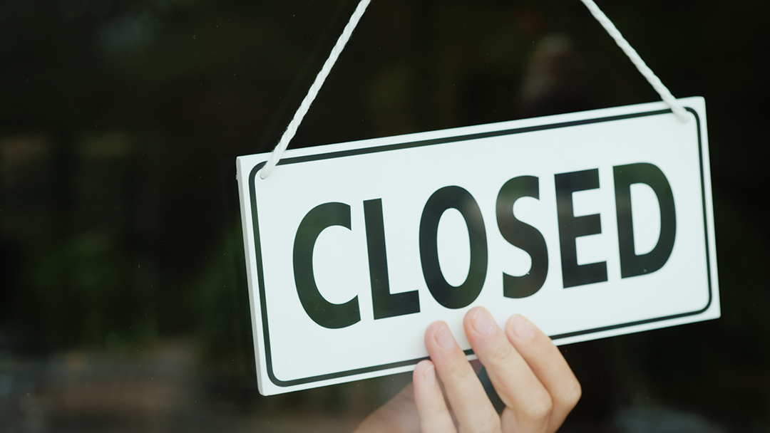 A hand holds a hanging “CLOSED” sign on a glass storefront, with a blurred interior.